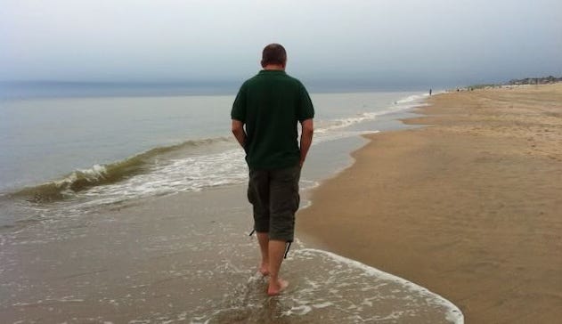 A man walks barefoot along a quiet beach, deep in thought, hands in his pockets, as gentle waves lap at the shore under a hazy sky.