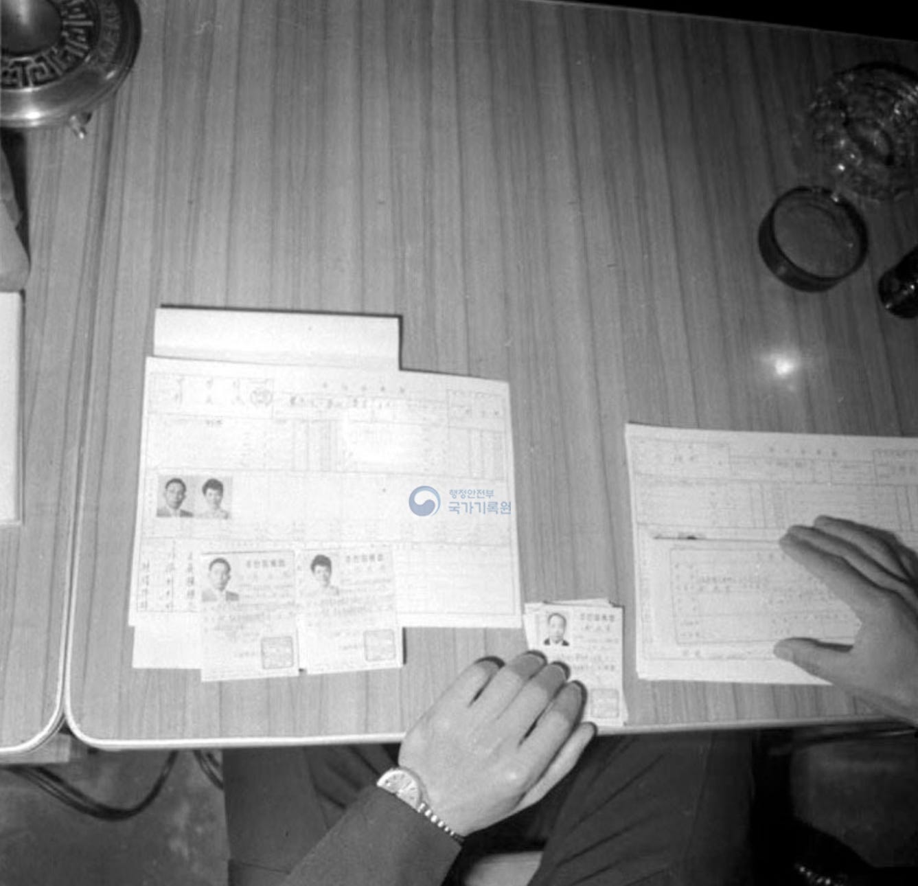 Black-and-white overhead photo of a desk with resident-registration forms laid out, small portrait photos attached, and several newly issued South Korean ID cards. An official’s hands sort the documents; a bell and glass ashtray sit at the corner. Watermark: National Archives of Korea.