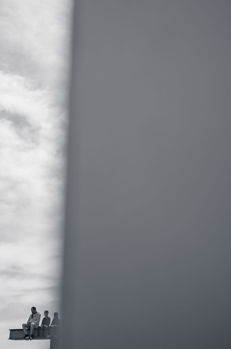 Three people seated on a narrow steel beam at Rockefeller Center, surrounded by bright sky, framed by a large expanse of negative space in black and white. Three people seated on a narrow steel beam at Rockefeller Center, surrounded by bright sky, framed by a large expanse of negative space in black and white.