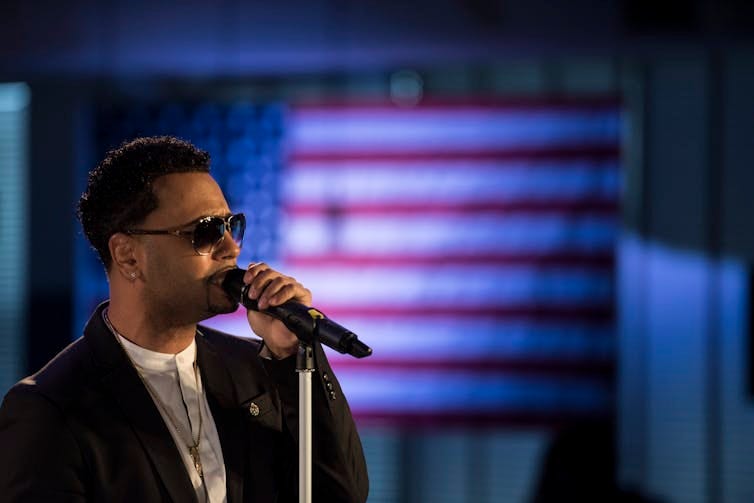 Man wearing suit and sunglasses sings into microphone with American flag hanging in background Man wearing suit and sunglasses sings into microphone with American flag hanging in background