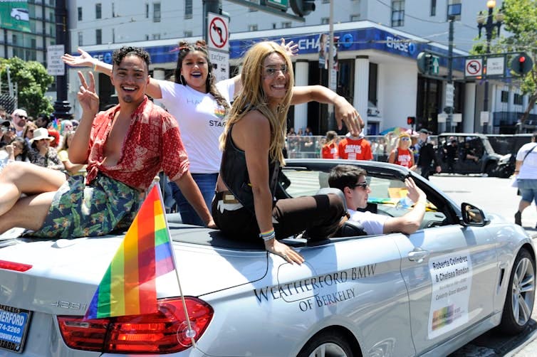 Three smiling LGBTQ Latinos ride along a city street in an open-top convertible with a rainbow flag