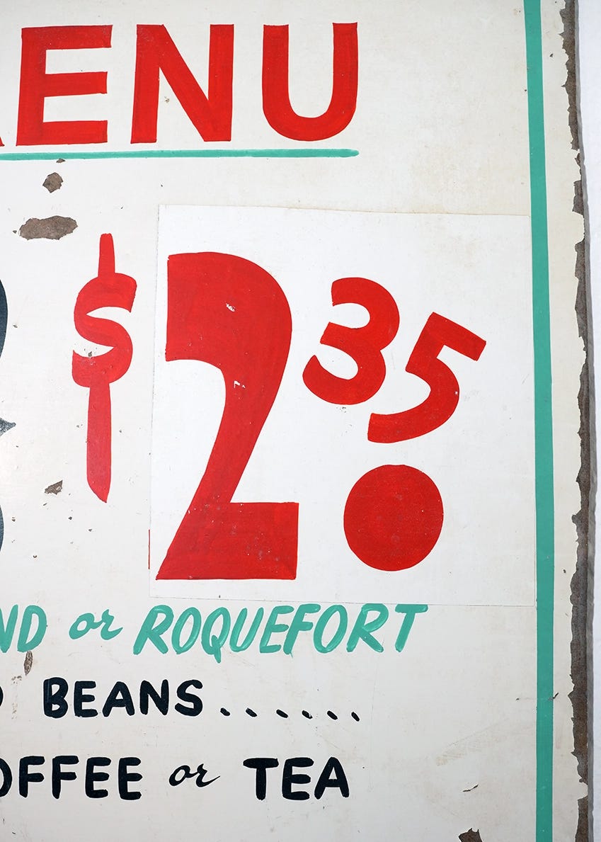 Close-up of a hand-painted restaurant menu board showing a $2.35 price in oversized red numerals, with menu items including Roquefort dressing, beans, and coffee or tea lettered below in teal and black.