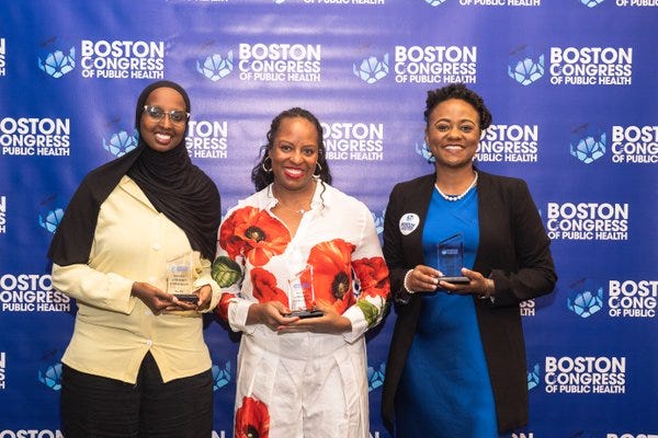 Three honorees with awards on the BCPH backdrop: a hijab-wearing honoree on the left, Miya Cain in a red poppy print in the middle, and a third honoree in royal blue on the right.