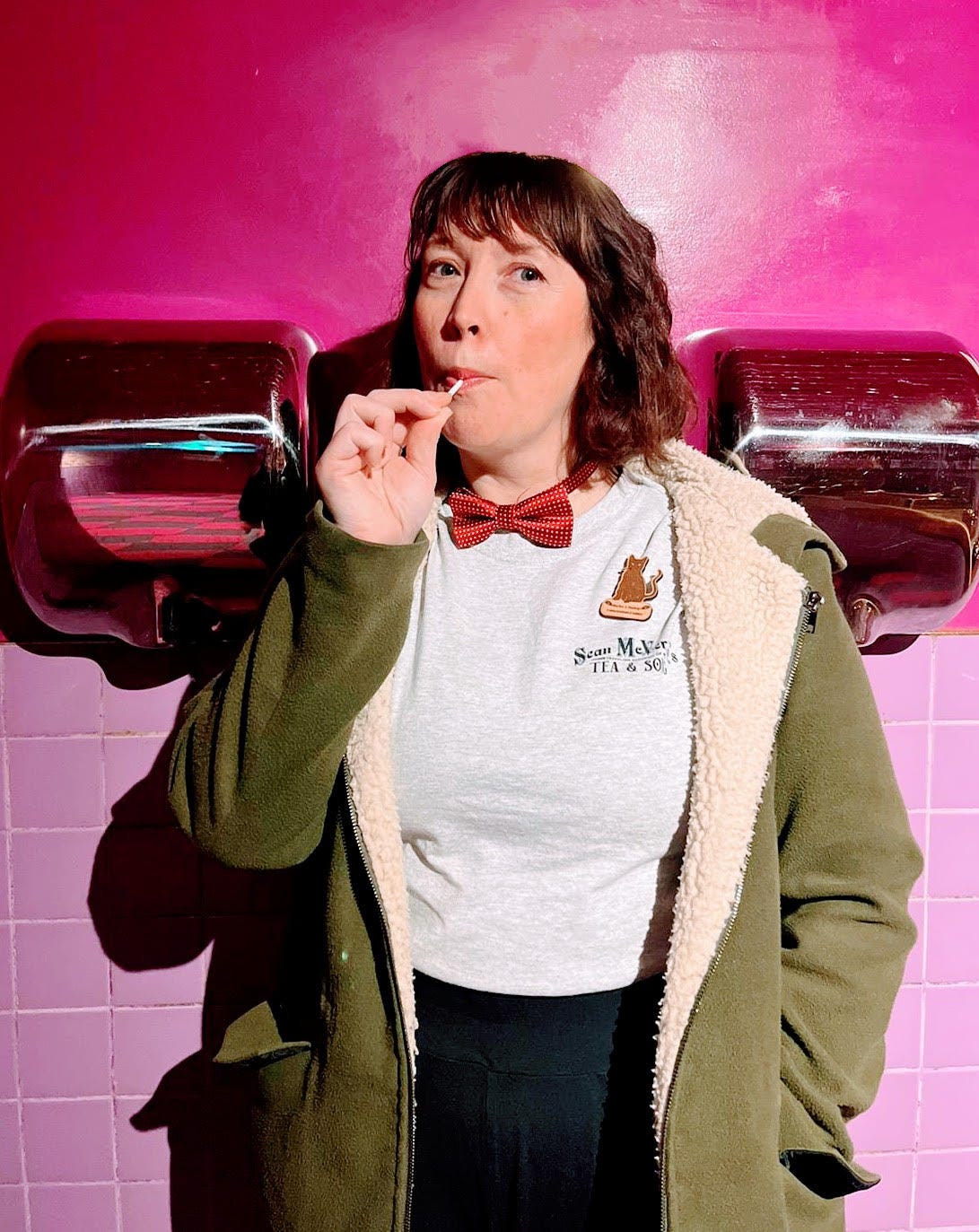Hayley is stood in between two Xlerator hand dryers against a bright pink wall. She is wearing a red bow tie, a big green coat and is sucking a lollipop. She looks weird but content.