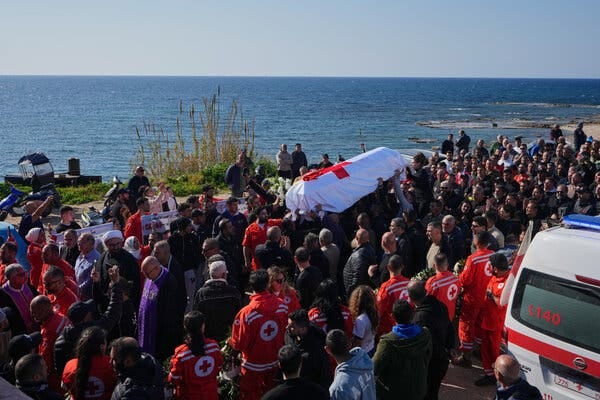 A crowd of people, many wearing Red Cross uniforms, lifts up a coffin during a funeral procession.