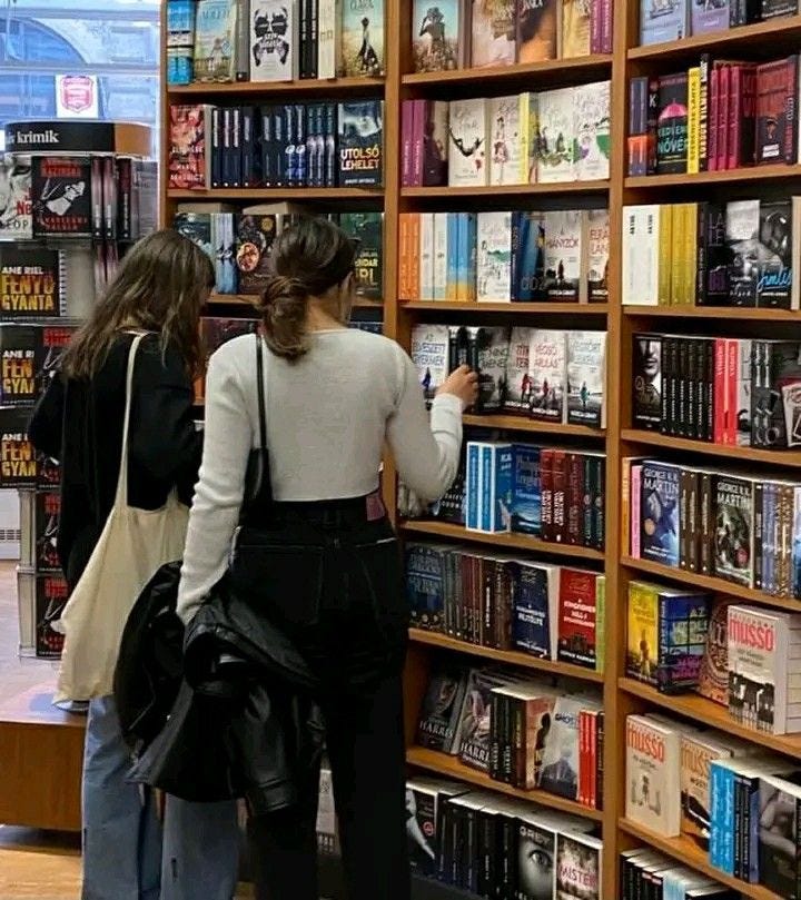 This may contain: two women looking at books in a book store with the shelves full of books behind them