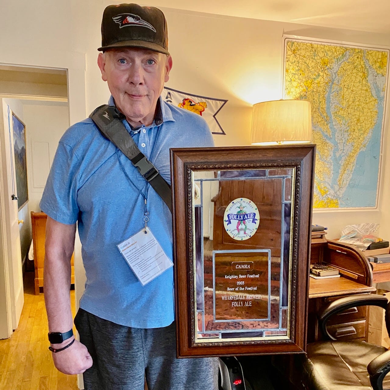 A man holding a large framed mirrored award plaque, standing in a home interior, a desk, lamp, and map visible on the wall behind him. 