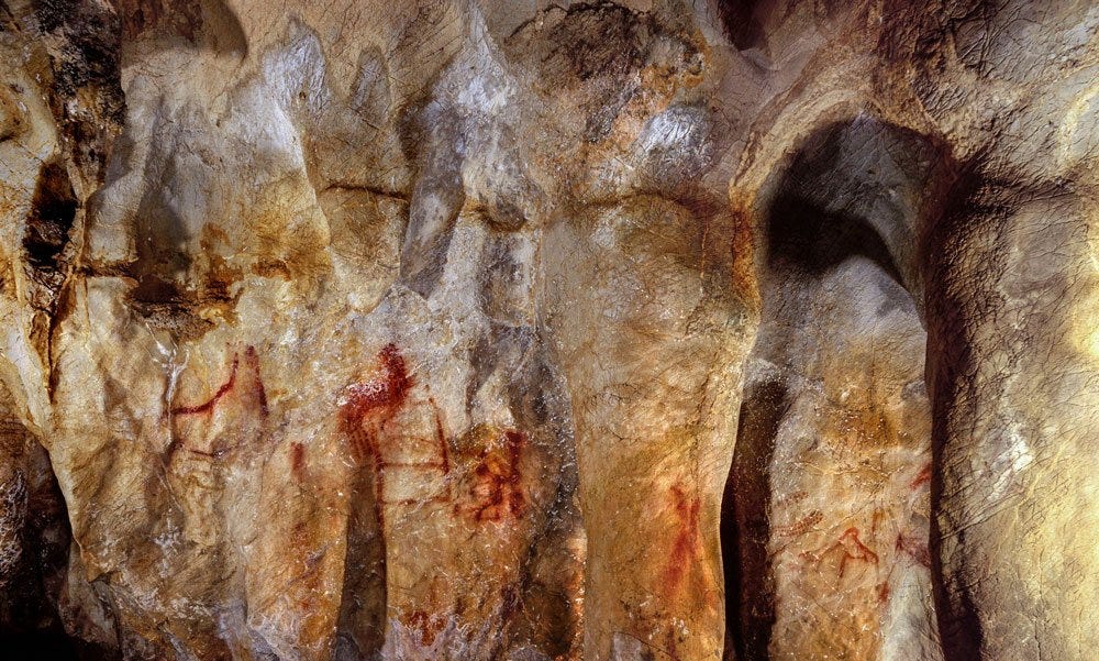 Verschiedene Zeichnungen in rotem Ocker auf unregelmäßigen Höhlenwänden, die prähistorische Kunsttechniken und frühe Ausdrucksformen des Menschen veranschaulichen.