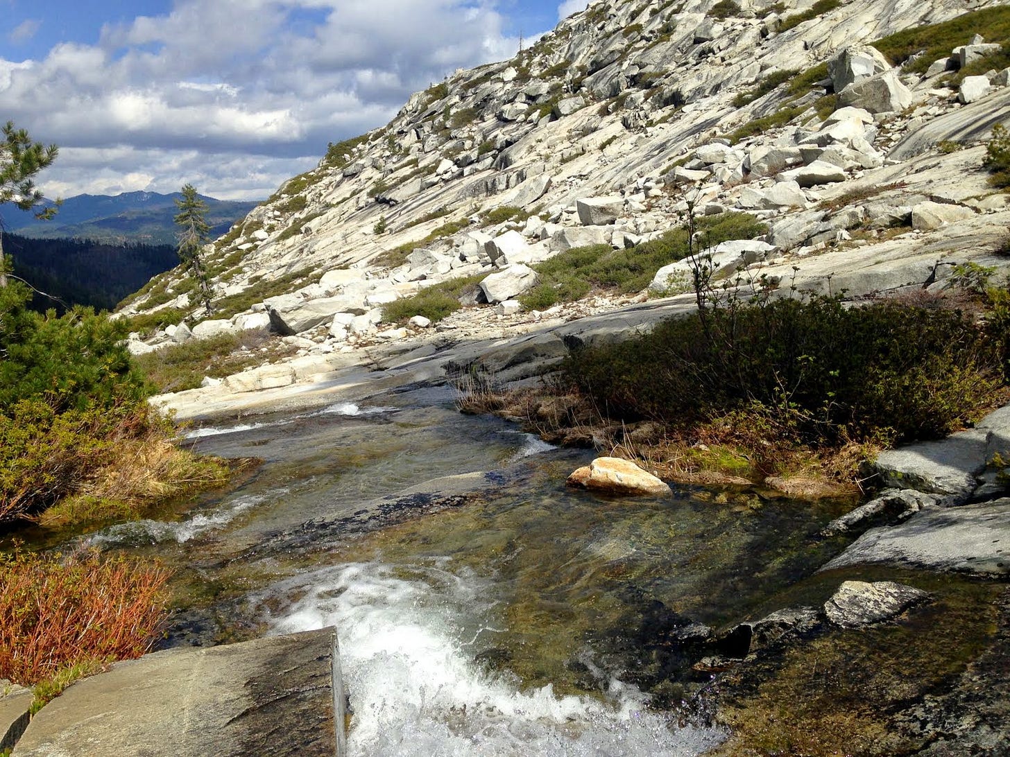 A shallow stream runs down the side of a mountain composed of blocky white rocks with mountains and evergreen trees off into the distance.