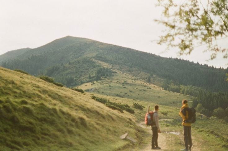 wo hikers with backpacks stand on a winding dirt trail, looking toward rolling green hills and a forested mountain under soft daylight.