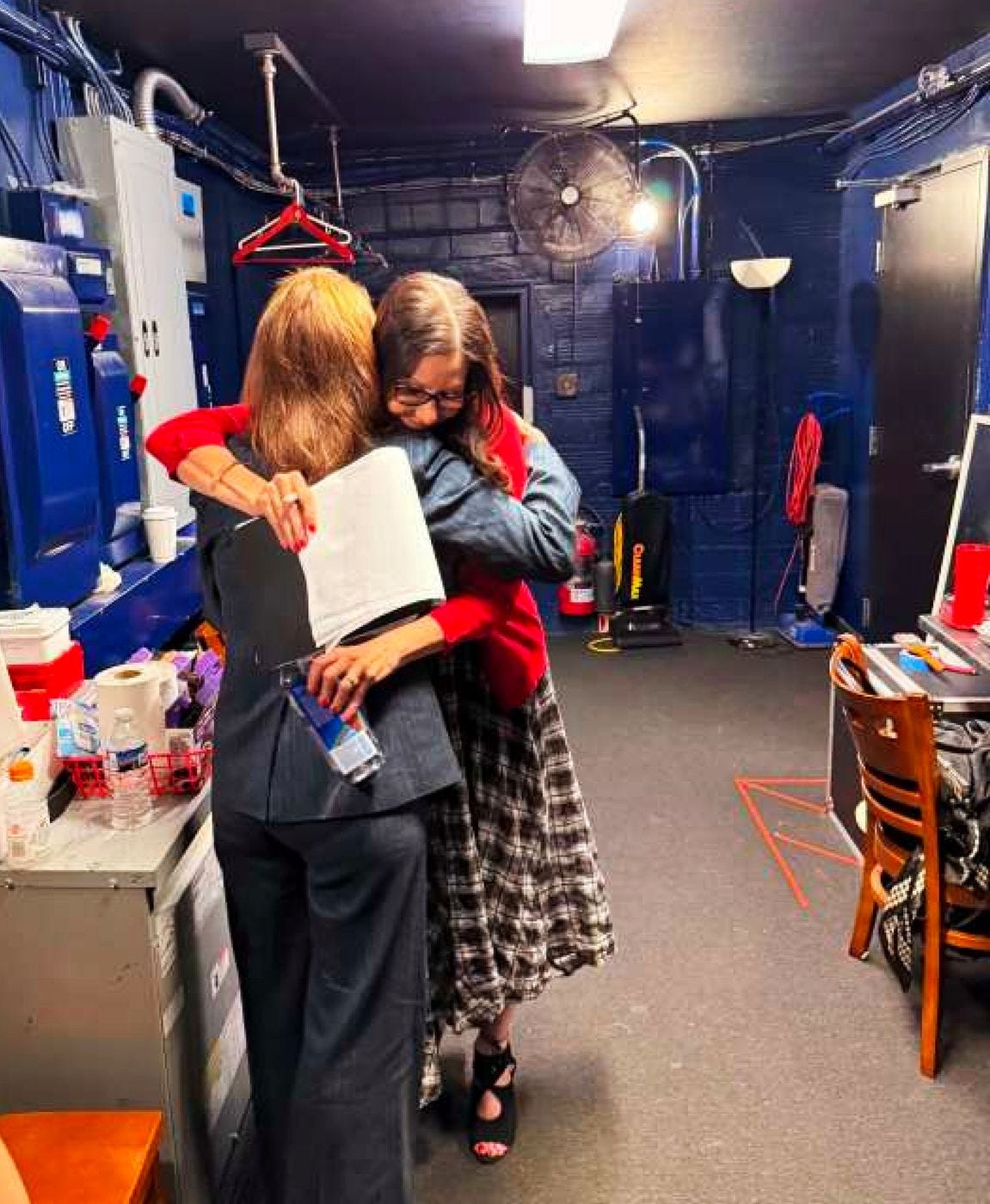 Two women embrace in a blue-painted backstage utility room lined with electrical panels, a commercial vacuum against the far wall. One still holds a clipboard against the other's back. Two women embrace in a blue-painted backstage utility room lined with electrical panels, a commercial vacuum against the far wall. One still holds a clipboard against the other's back.