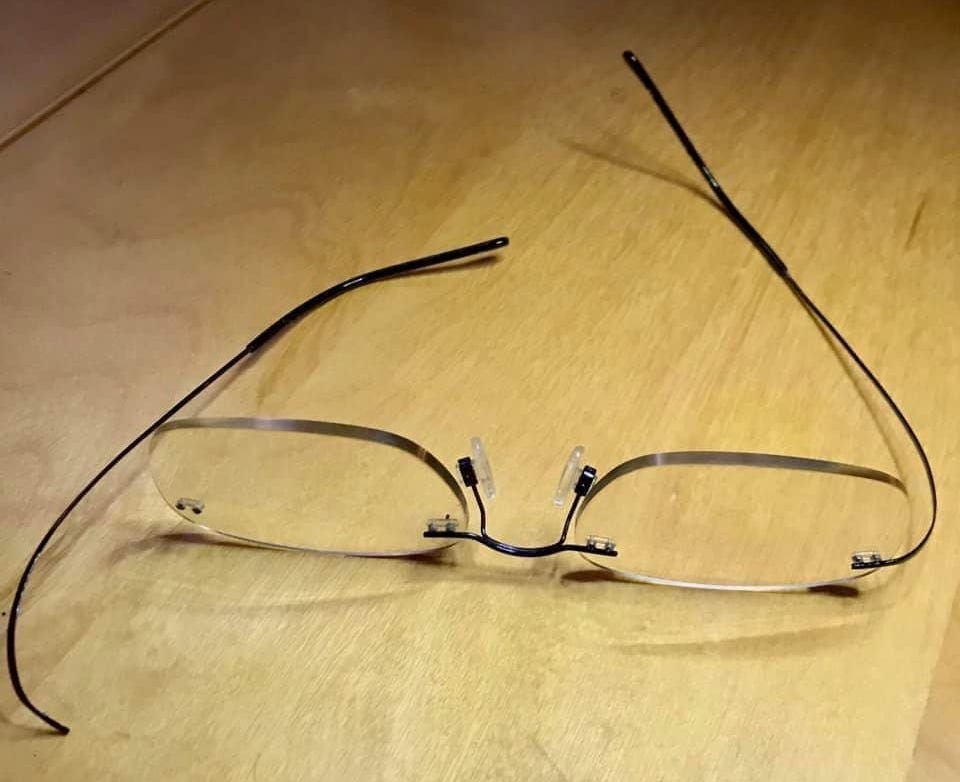 a photograph of a pair of rimless glasses with the left arm broken, on a pale wood surface