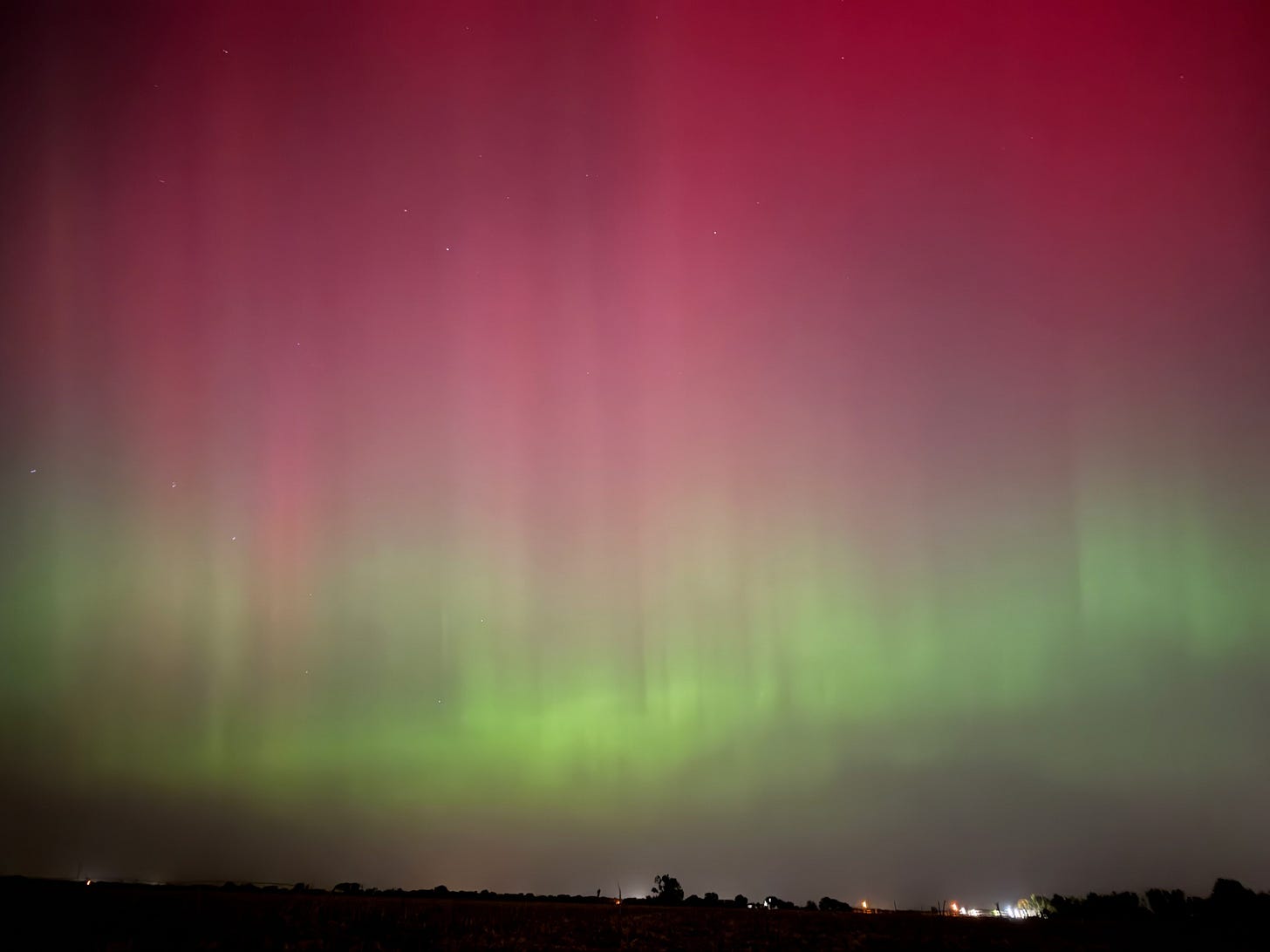 Pillared and wavy aurora borealis, reddish pink higher in the sky and lime green lower to the ground, over a dark, silhouetted flat horizon.