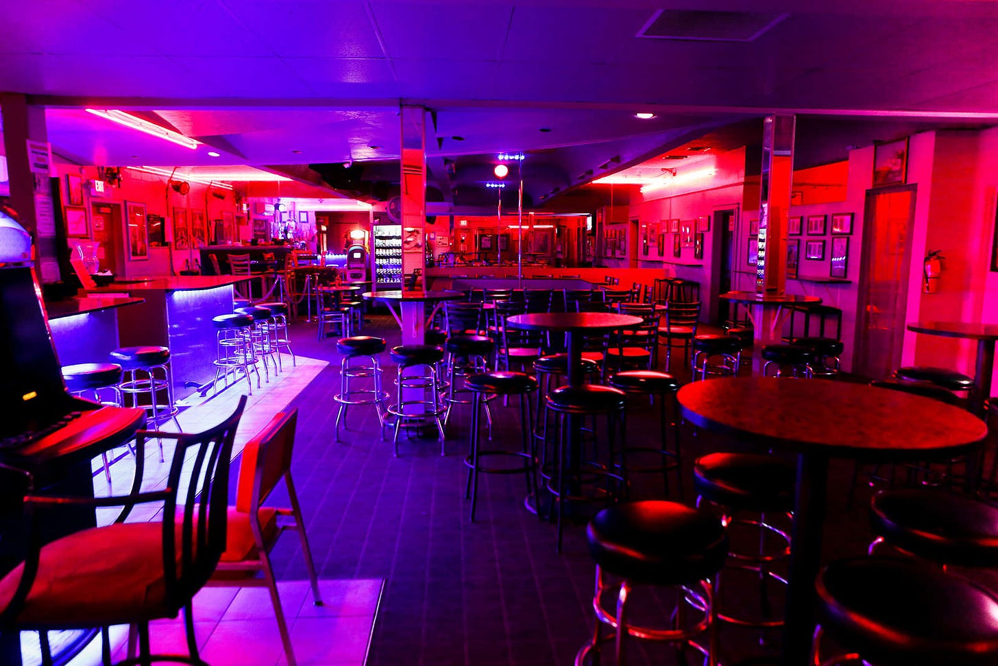 Interior of a dimly lit club with empty tables and chairs bathed in pink and purple neon lighting, creating a moody, atmospheric scene that evokes contemplation and introspection.