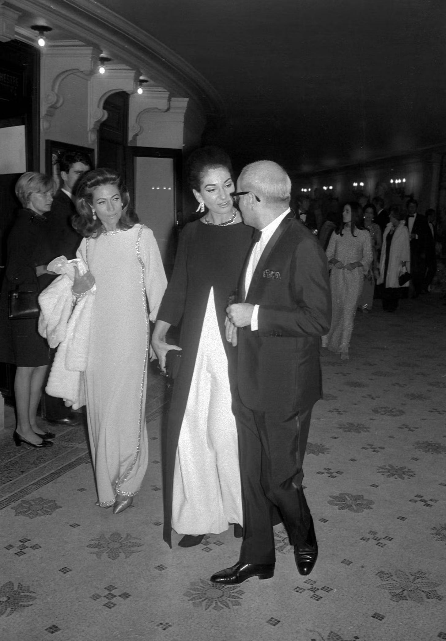 A woman stands flanked by a woman and a man while wearing a dark, spliced overlay with long sleeves on top of a flowing white underdress.