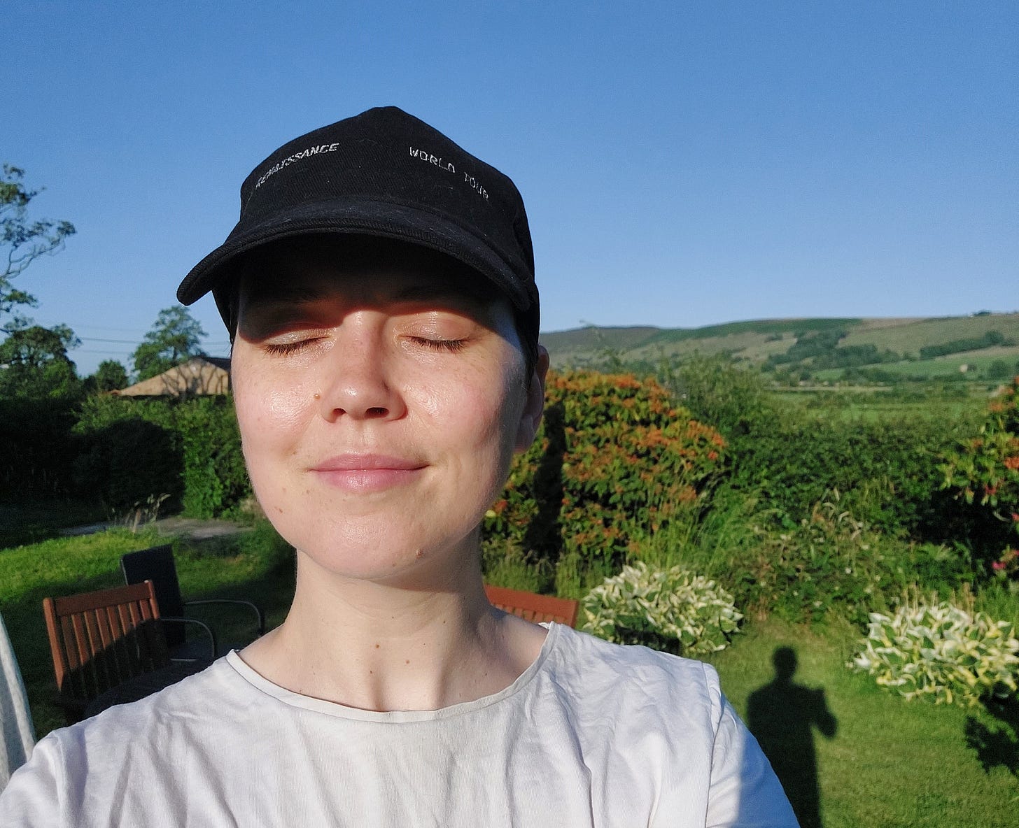 Head and shoulders of Janelle (white woman wearing a black cap) with her eyes closed and the sun shining on her face. Behind her blue sky and green rolling fields