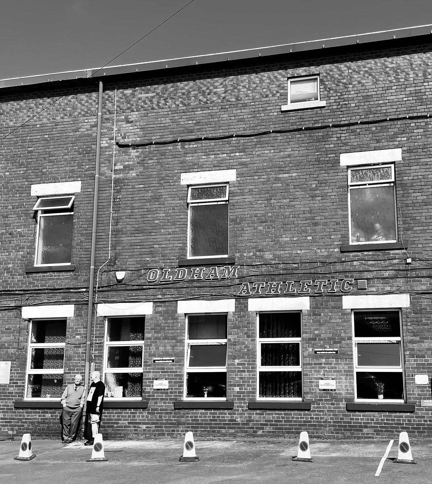 A black-and-white image of the exterior of the old stand - very 1970s style lettering on the wall reads 'Oldham Athletic'.