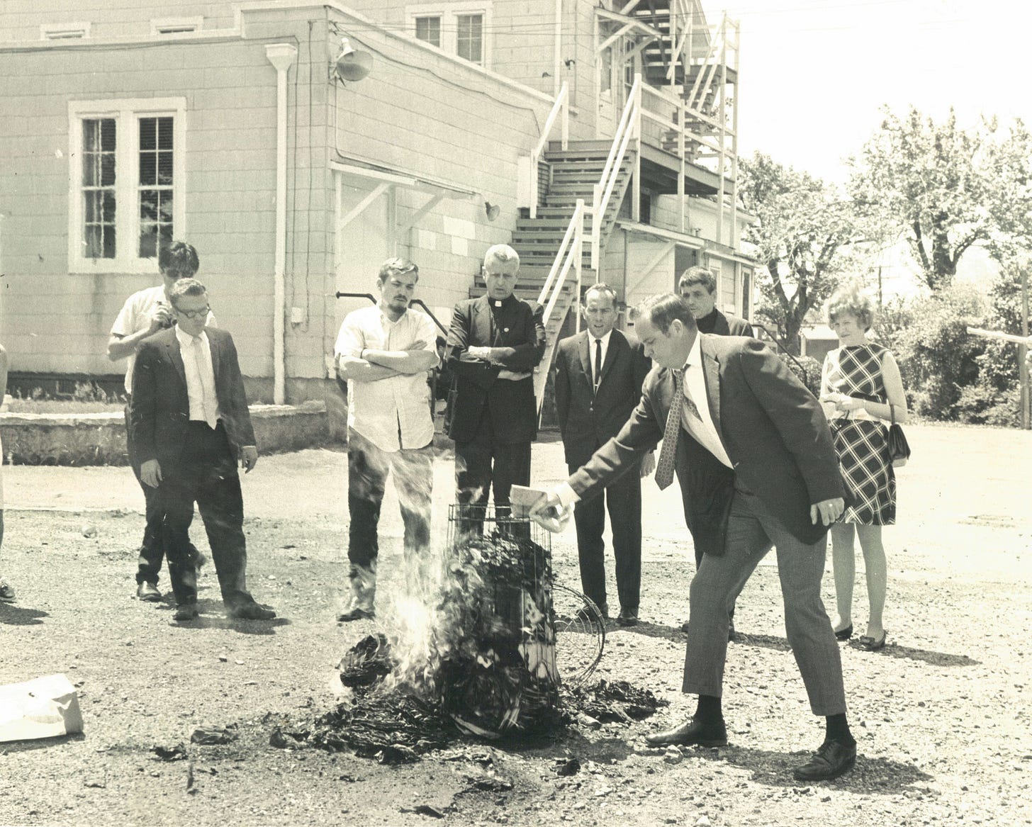 A group of people, mostly men in suits but including at least one woman, stand outside a building where there is a fire. They are burning stolen draft cards.