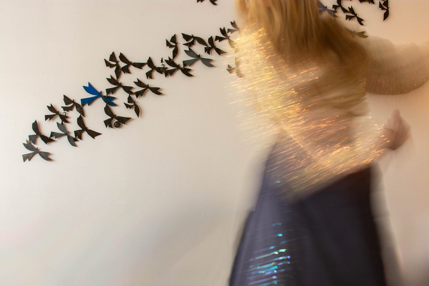 photo of bird sculpture on a wall with a woman in a sparkly blue dress in front of it - she is blurred in motion and her blond hair is also in motion