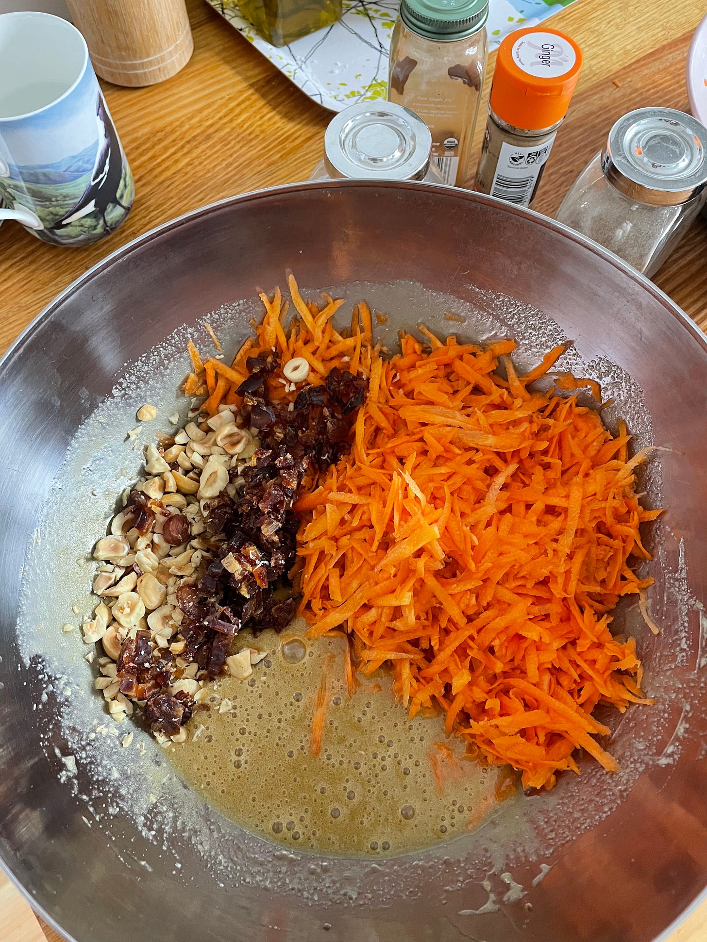 Silver bowl with carrot cake ingredients ready to mix, including chopped medjool dates, hazelnuts and grated carrot.
