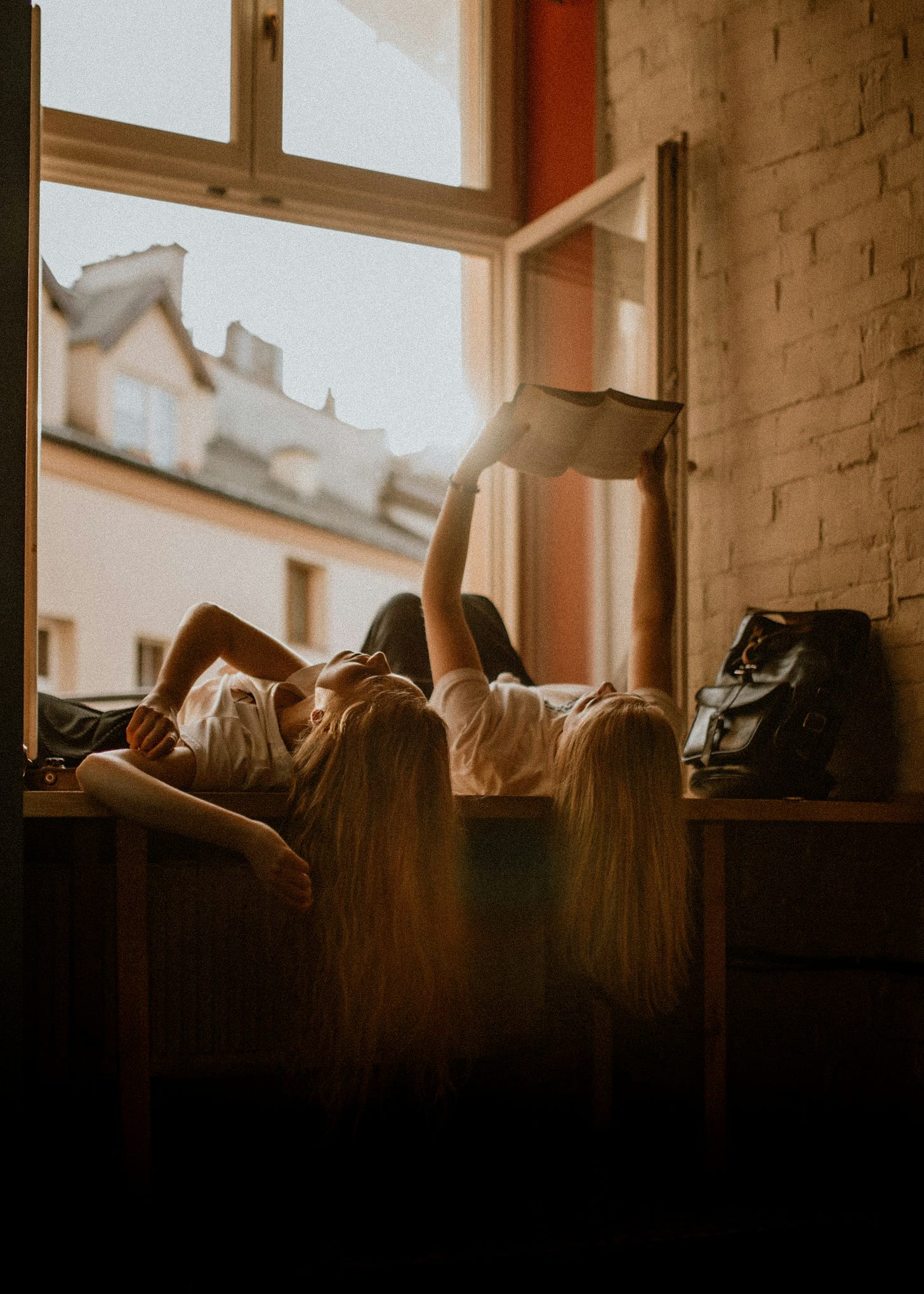 Two girls lying in front of an open window, one holding an open book straight up over her face to read.