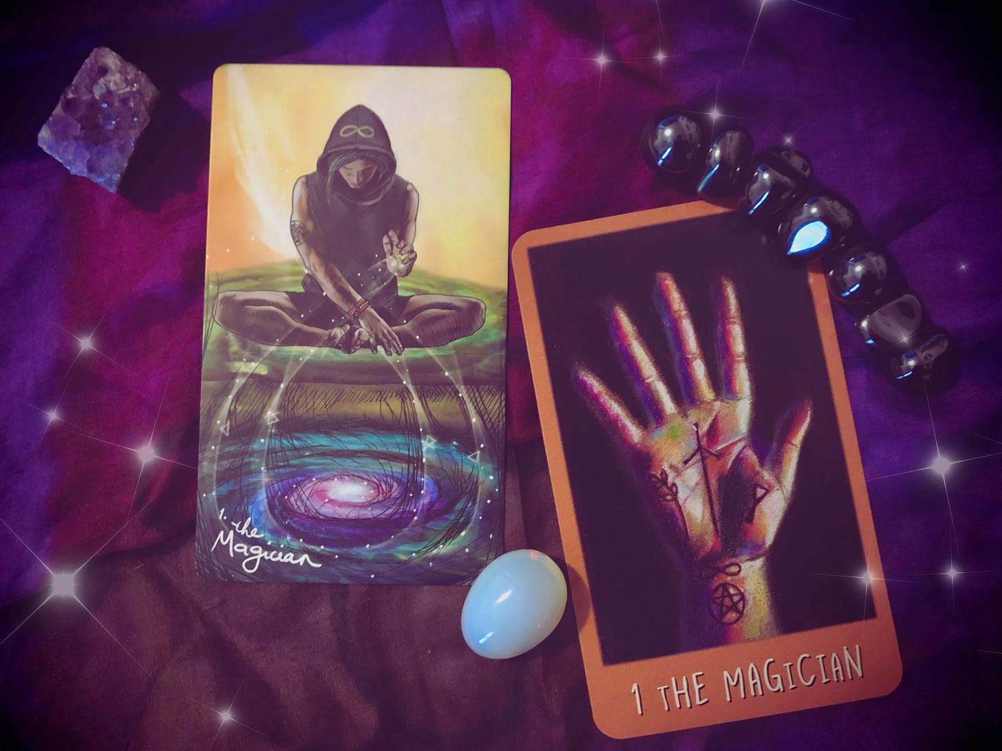 Two Magician tarot cards from different decks on a purple cloth, surrounded by crystals — an amethyst cluster, a black bracelet, and an opalite stone. The left card shows a hooded figure sitting cross-legged, channeling energy into a swirling galaxy pool below. The right card shows an open palm with mystical symbols (a cross, a triangle, a pentacle).