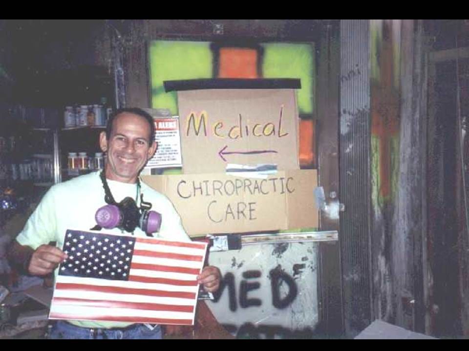 Smiling man with a respirator around his neck holds an American flag beside handmade Medical and Chiropractic Care signs