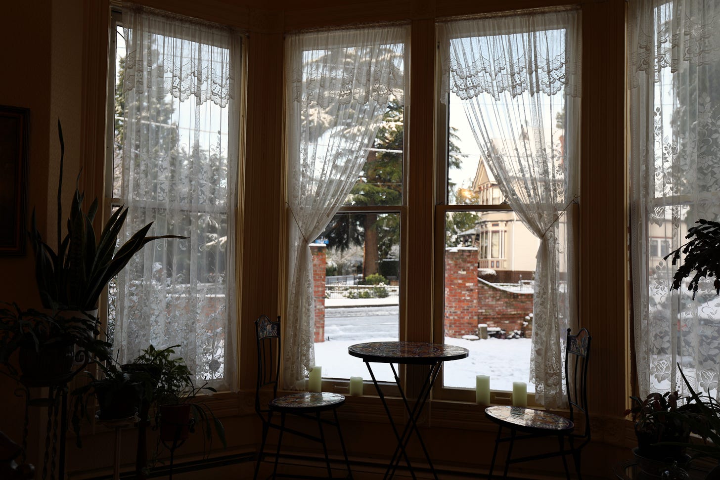 Interior view of a Victorian parlor with lace curtains, small table and chairs, and a snowy street visible through tall windows.
