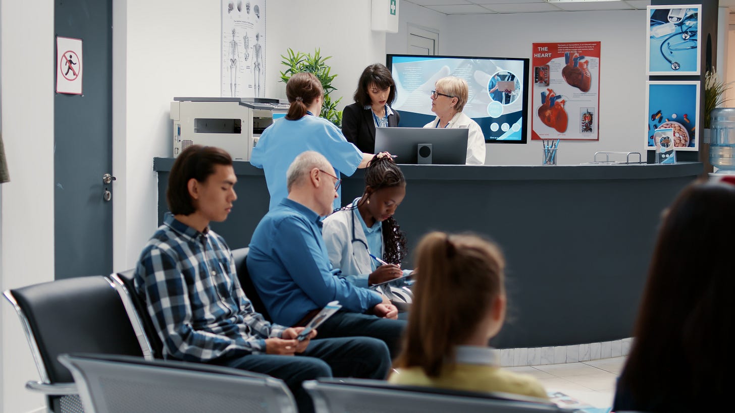 Pazienti seduti nella sala d'attesa di una clinica medica, mentre due receptionist e un medico parlano al bancone della reception. Poster anatomici e monitor digitali decorano l'ambiente.
