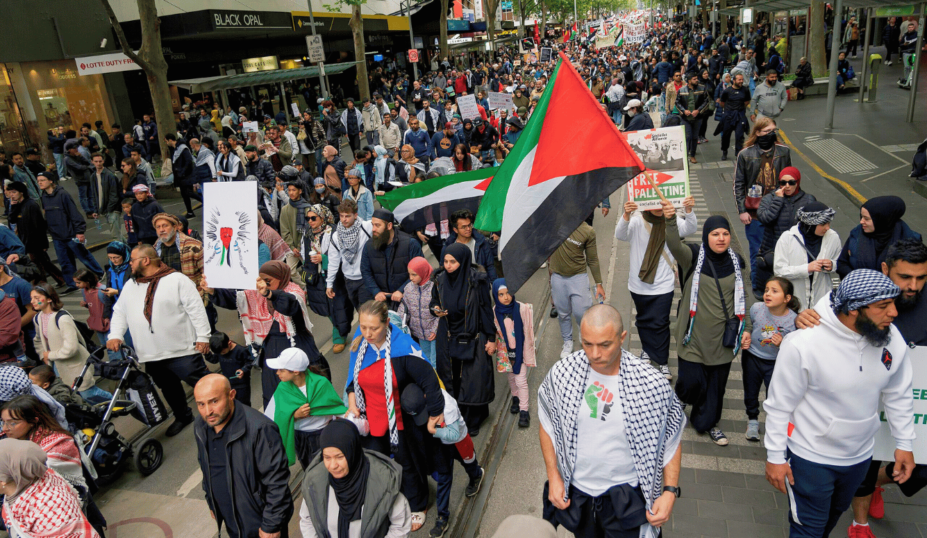 Large crowd marching along a city street during a pro-Palestinian protest, with Palestinian flags and placards visible.