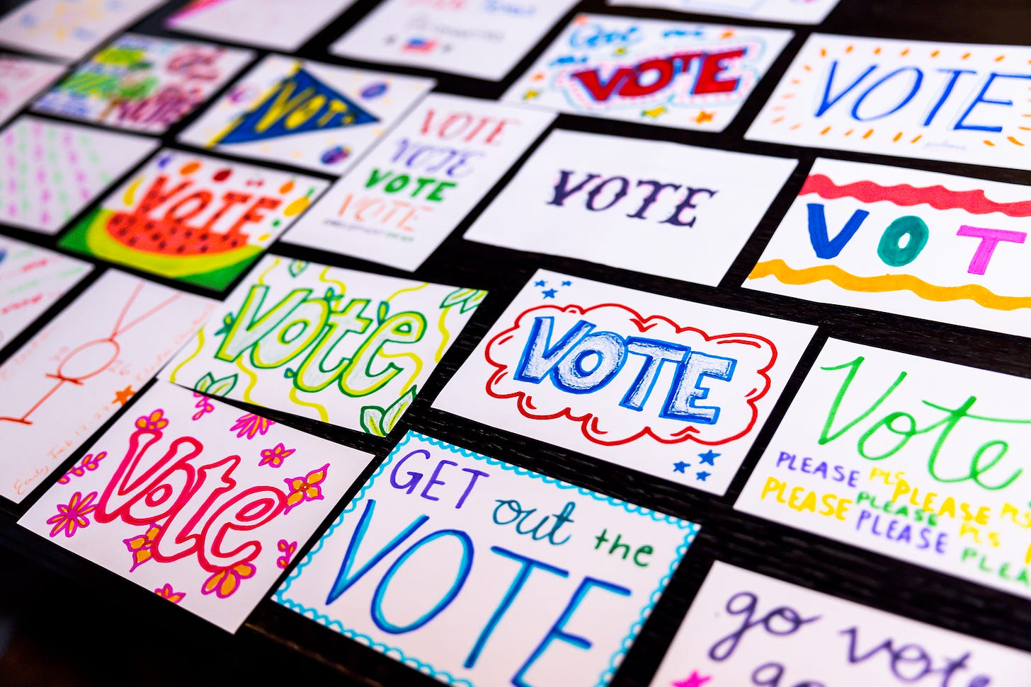 Multiple hand-made postcards laid out on a black table. Some say "vote" while others say "Get out the vote" and "please vote." All using variety colors.