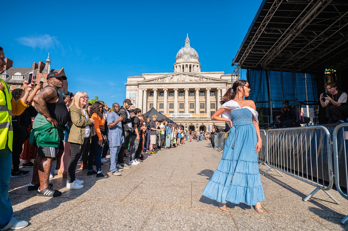 Nottingham's Old Market Square with a crowd of people to the left, a woman to the right posing in a blue dress, and a large stone building and a blue sky in the background Nottingham's Old Market Square with a crowd of people to the left, a woman to the right posing in a blue dress, and a large stone building and a blue sky in the background