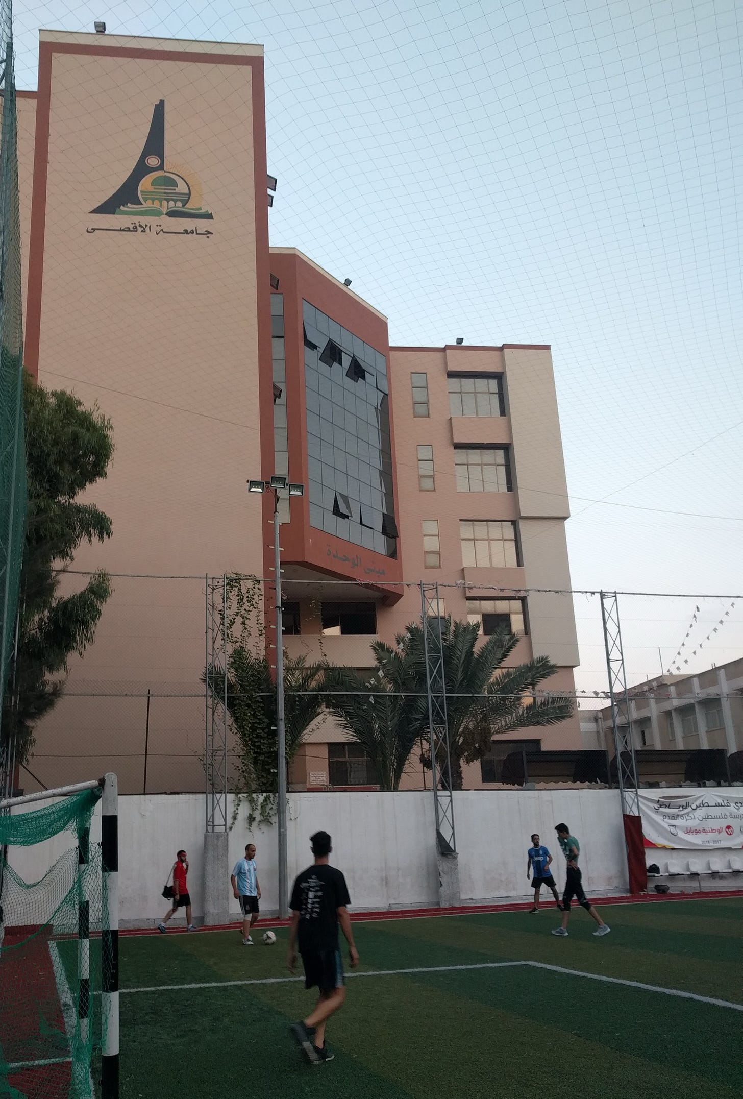 Men play soccer on a fenced-in turf field with a large building behind them