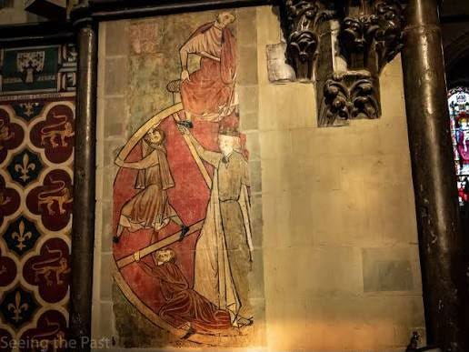 A faded medieval wall painting inside a church, showing robed figures positioned around a large circular form beside ornate stonework. A faded medieval wall painting inside a church, showing robed figures positioned around a large circular form beside ornate stonework.