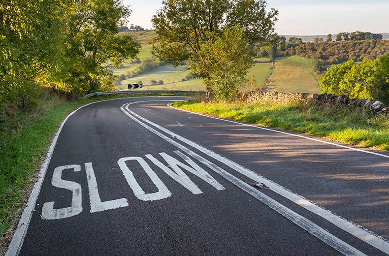 the word slow is painted on the right hand side of a road in big white letters. The tarmac road is curving through green countryside. the word slow is painted on the right hand side of a road in big white letters. The tarmac road is curving through green countryside.