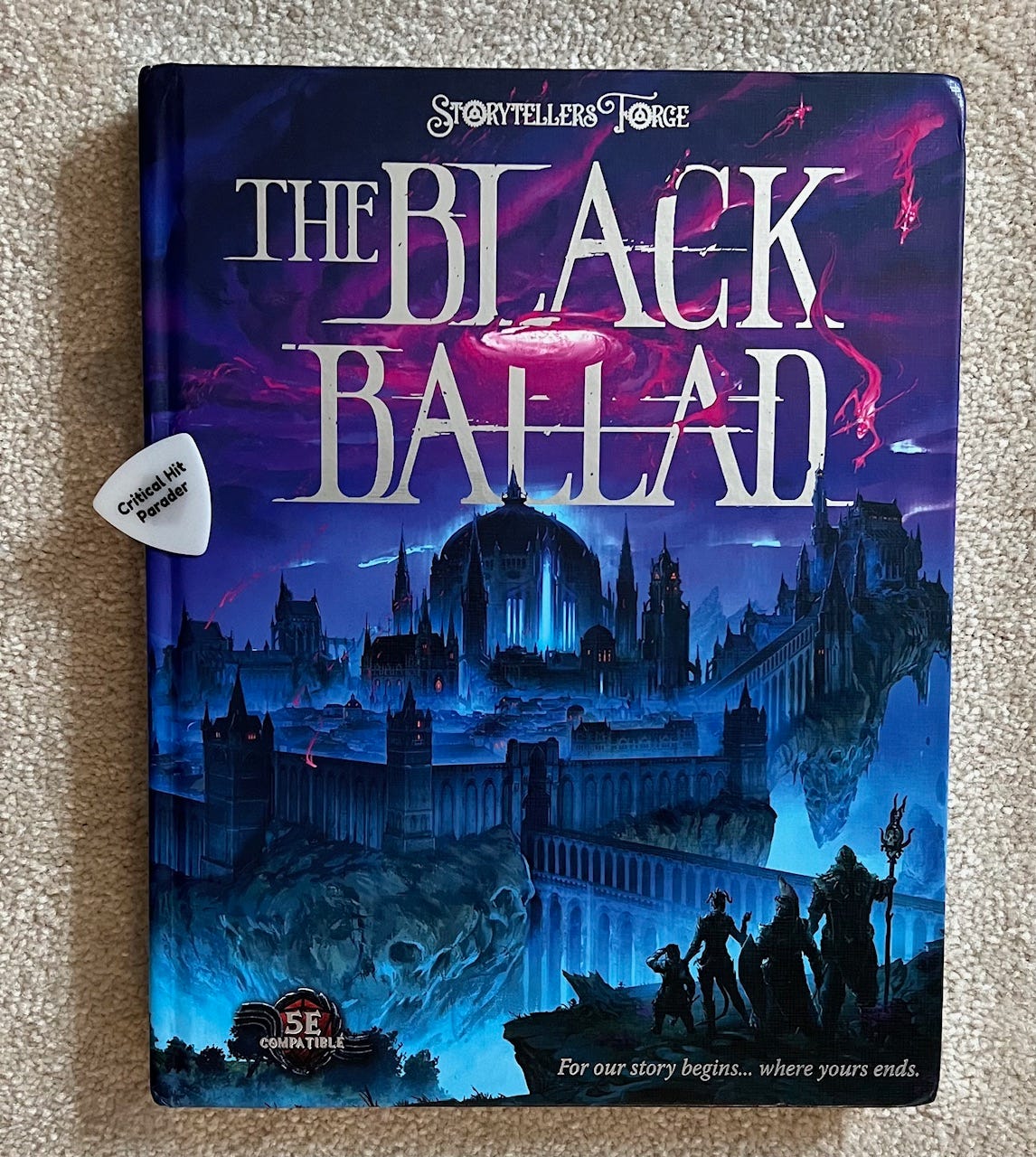 A photo of The Black Ballad TTRPG book. It features artwork of a shadowed RPG party in front of a fantasy city at night. The primary colors are black and blue. A white Critical Hit Parader guitar pick is laying on top of the book.