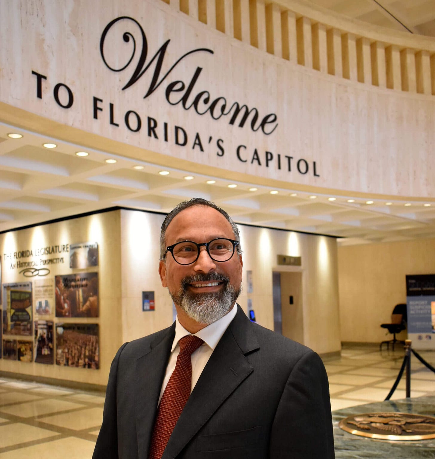 Ramon Maury standing in Florida’s Capitol building, smiling in a dark suit, white shirt, and red tie, with a large “Welcome to Florida’s Capitol” sign above him.