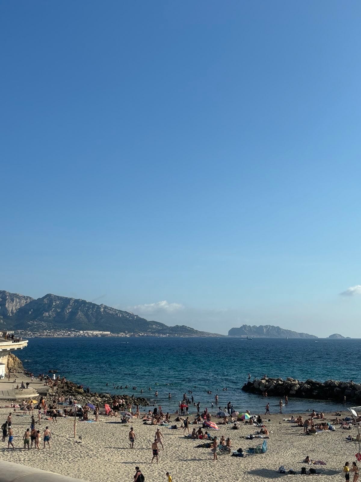 A photo of Plague de Prophet from an upper railing. You can see the beach below with its various enjoyers milling about or wading. You can see a part of the rock formation off the coast that forms tide pools and a shallow pool safe from the open water. In the background, behind the rich teal water, are jagged green and low mountains. The light blue ombre sky takes up most of the image.