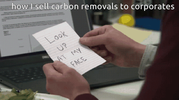 Animated GIF captioned "how I sell carbon removals to corporates". A hand slides a handwritten note reading "LOOK UP AT MY FACE" across a desk. The man at the desk, in a maroon sweater, looks up at the man standing over him, who says "You gotta give."