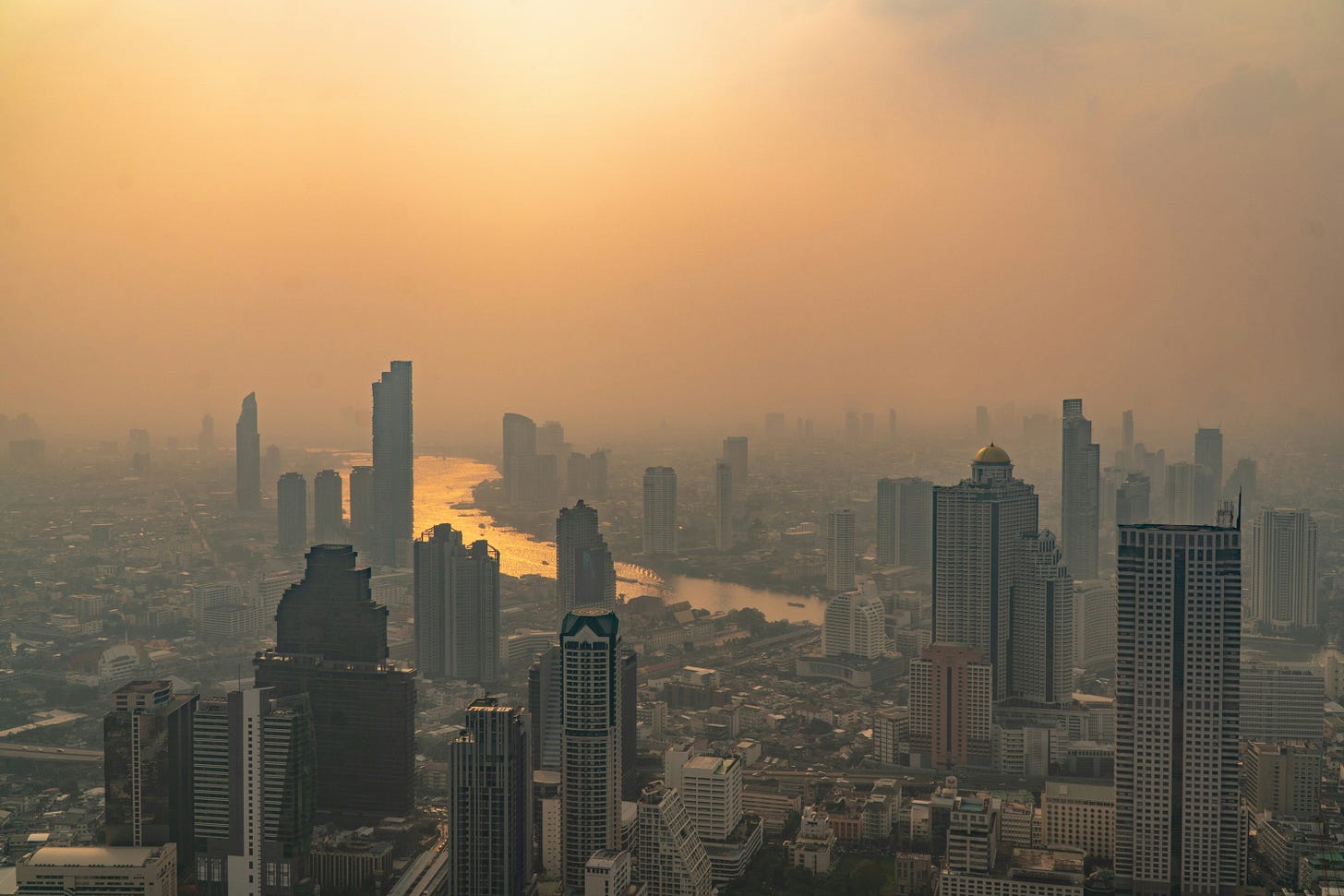 An aerial, high-angle photograph of Bangkok, Thailand, showing a sprawling metropolitan skyline, featuring numerous skyscrapers and a winding river, heavily blanketed under a thick, murky layer of orange-brown smog and toxic haze. The oppressive atmospheric conditions visually ground the newsletter's report on the escalating dual climate emergency of extreme humid heatwaves and severe wildfire air pollution affecting major Southeast Asian cities like Bangkok. An aerial, high-angle photograph of Bangkok, Thailand, showing a sprawling metropolitan skyline, featuring numerous skyscrapers and a winding river, heavily blanketed under a thick, murky layer of orange-brown smog and toxic haze. The oppressive atmospheric conditions visually ground the newsletter's report on the escalating dual climate emergency of extreme humid heatwaves and severe wildfire air pollution affecting major Southeast Asian cities like Bangkok.