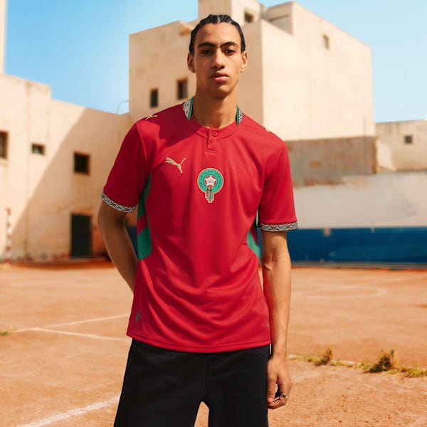 Morocco Home Men's Soccer Jersey, Fast Red-Victory Gold, extralargeA model wearing Morocco’s 2026 Puma home jersey in deep red with green side panels, gold Puma logo, and Moroccan crest, standing outdoors against a pale architectural backdrop.