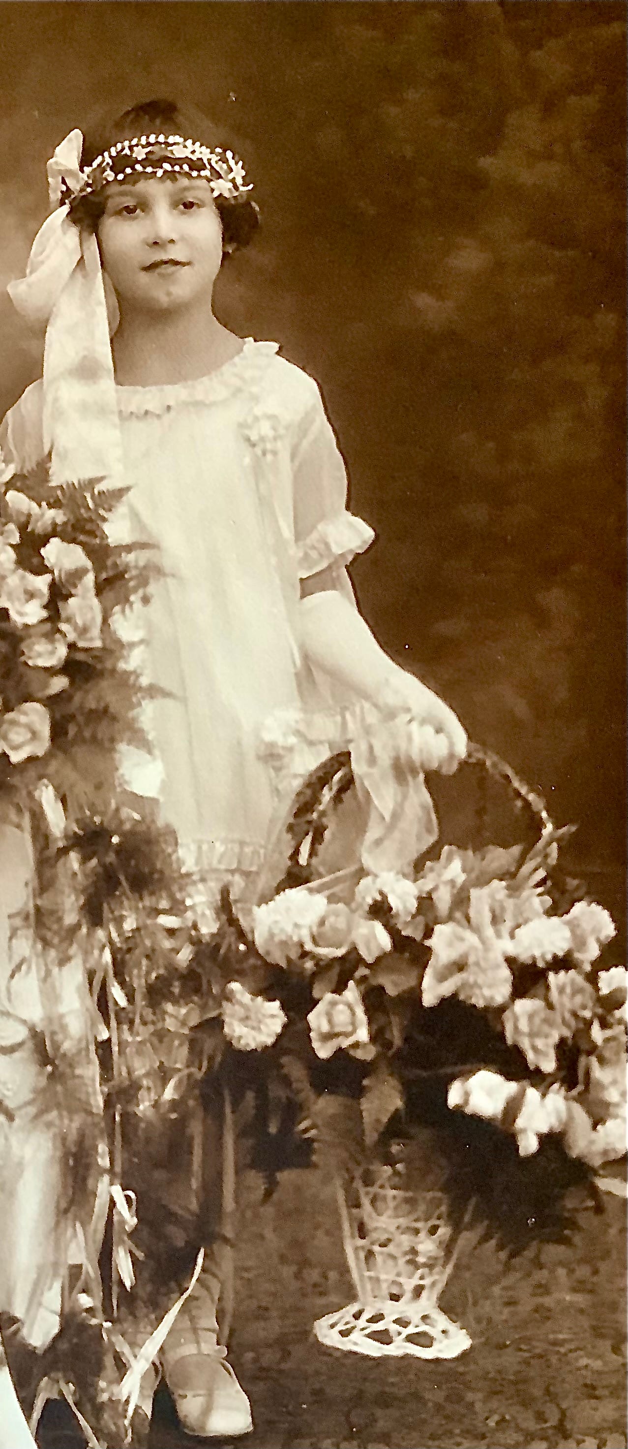 A young Italian girl in a lovely white dress, white stockings and shoes. She has a headpiece on and holds the handle of a basket with white roses flowing out of it. She's in a wedding.