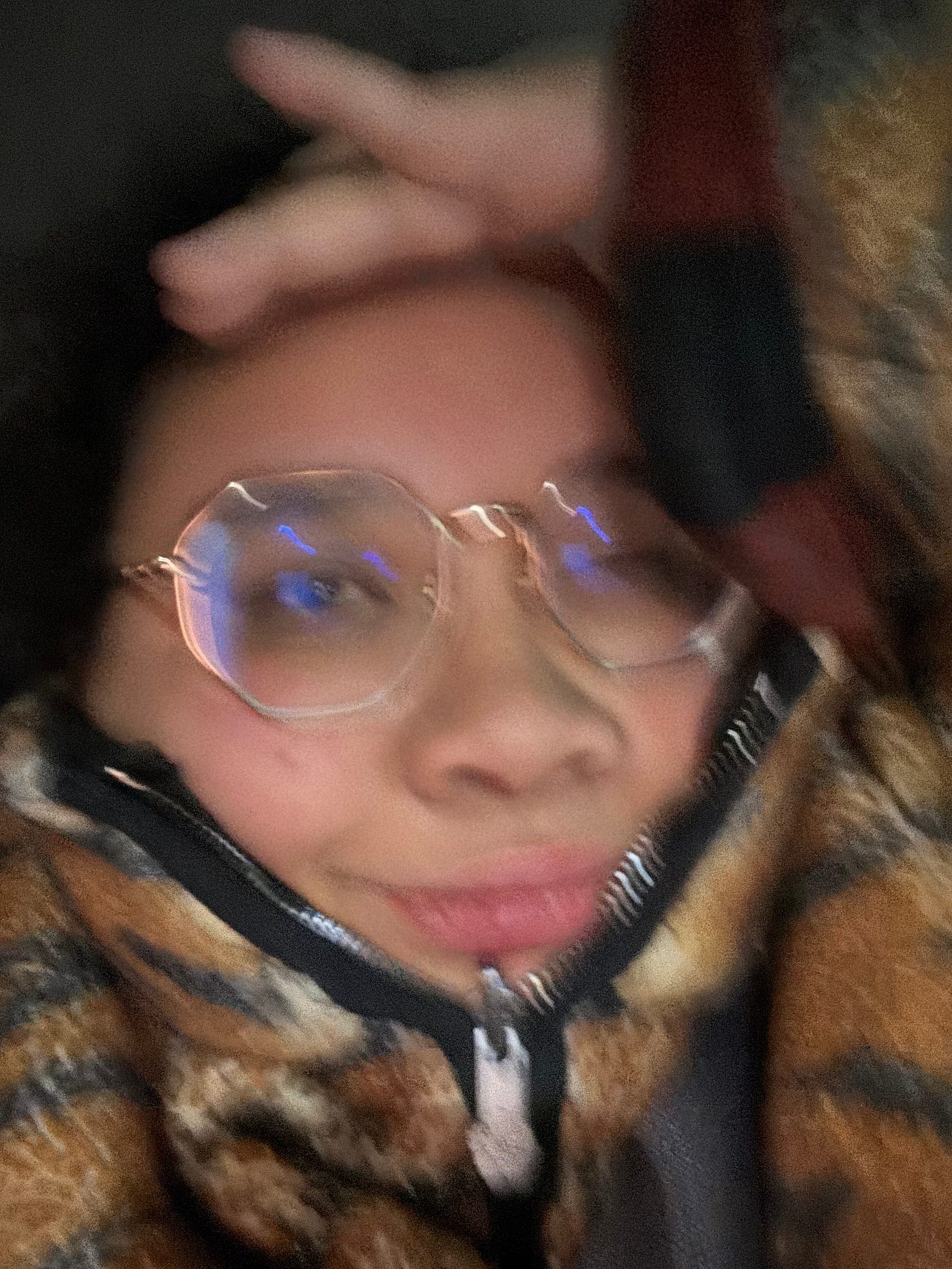 A blurry close-up selfie of a person wearing round glasses and a patterned zip-up jacket with a high collar covering most of their neck. Their lips are slightly pursed, and their expression appears relaxed or amused. One hand is resting on their forehead. The lighting is dim, with a soft glow reflecting off their glasses. The background is indistinct due to the focus and motion blur. A blurry close-up selfie of a person wearing round glasses and a patterned zip-up jacket with a high collar covering most of their neck. Their lips are slightly pursed, and their expression appears relaxed or amused. One hand is resting on their forehead. The lighting is dim, with a soft glow reflecting off their glasses. The background is indistinct due to the focus and motion blur.