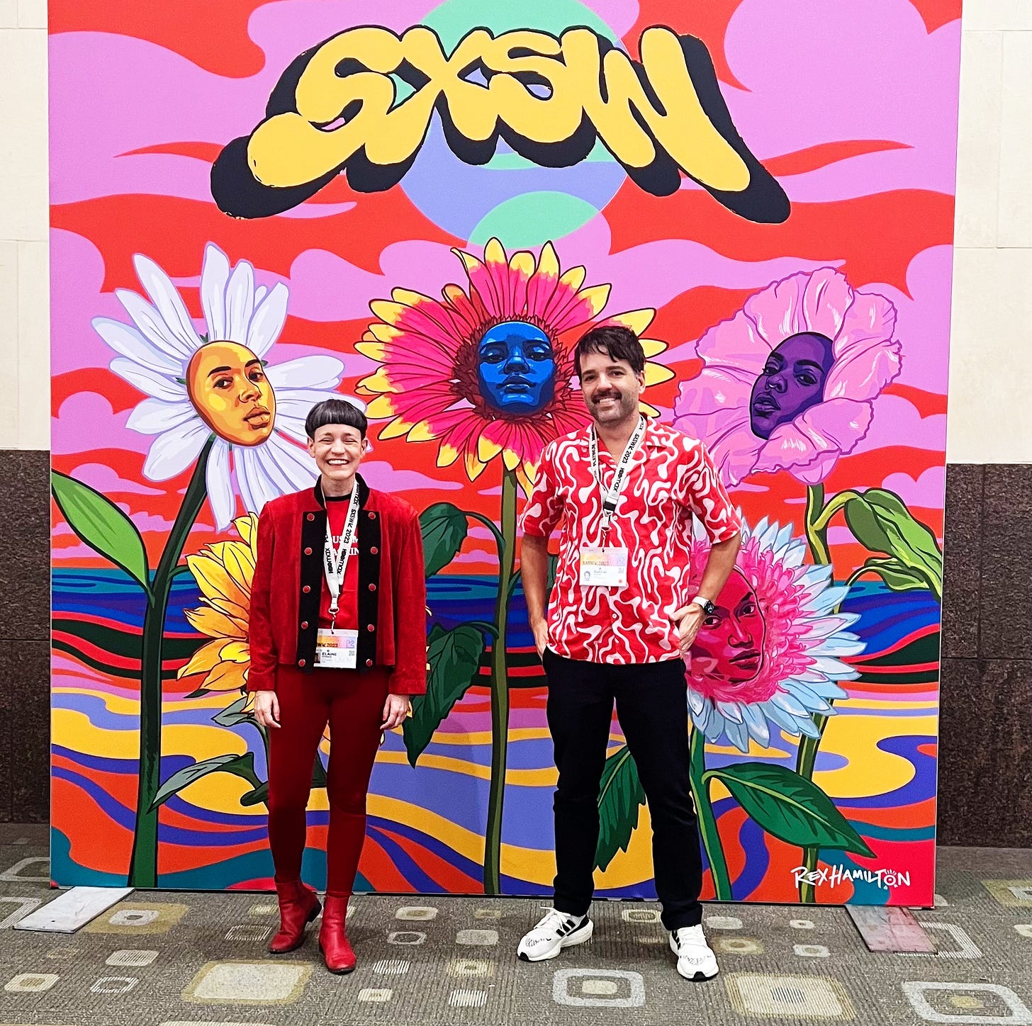 Placa do SXSW com 3 flores grandes desenhadas com rostos de mulheres no meio. Na frente, mulher magra, toda vestida de vermelho de calça e jaqueta, cabelo curto. Homem jovem de bigode ao lado sorrindo, vestindo camisa vermelha e branca, calça preta e tênis branco.