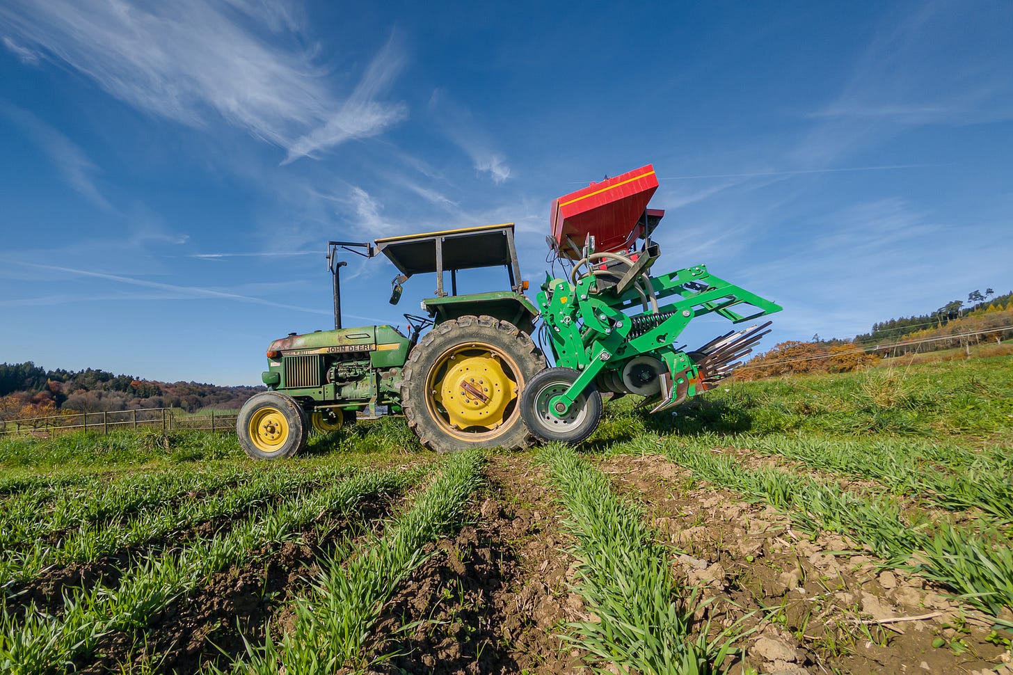 Ein historischer John-Deere-Traktor mit offenem Fahrersitz und Sonnendach steht auf einem Acker mit Reihen junger Pflanzen. Am Heck ist ein modernes, grün lackiertes Anbaugerät mit rotem Behälter befestigt, das einen deutlichen Kontrast zwischen alter Zugmaschine und neuer Technik bildet. Die Aufnahme ist aus tiefer Perspektive gemacht und zeigt im Hintergrund eine sanft hügelige Landschaft mit Waldrand und Herbstfarben. Ein historischer John-Deere-Traktor mit offenem Fahrersitz und Sonnendach steht auf einem Acker mit Reihen junger Pflanzen. Am Heck ist ein modernes, grün lackiertes Anbaugerät mit rotem Behälter befestigt, das einen deutlichen Kontrast zwischen alter Zugmaschine und neuer Technik bildet. Die Aufnahme ist aus tiefer Perspektive gemacht und zeigt im Hintergrund eine sanft hügelige Landschaft mit Waldrand und Herbstfarben.