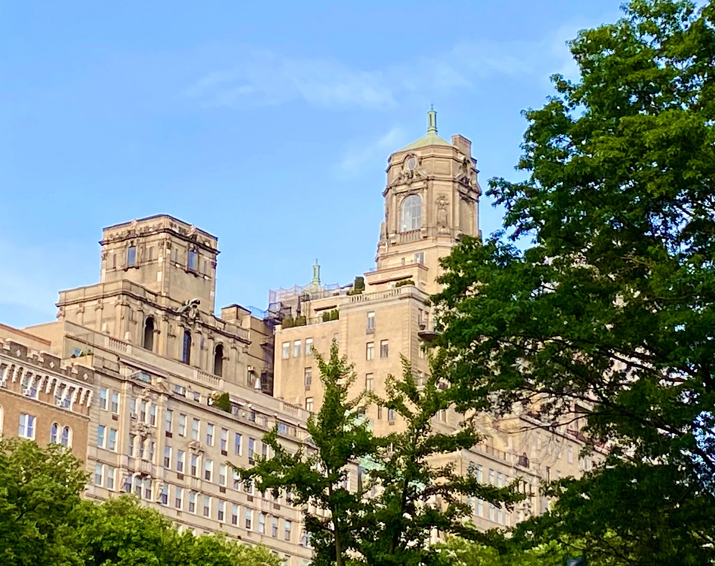 An ornate coop building against a blue sky with green trees in the foreground. Close to the center of the frame is an elaborate octagonal structure with a large window and a small round window above it.