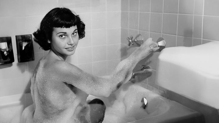 A four-image grid of vintage black-and-white photographs connected to Bunny Yeager: the top-left shows Yeager holding a camera while photographing a nude model lounging on the deck of a boat; the top-right shows a nude woman posing in a dramatic stance on an empty beach; the bottom-left shows a nude woman playfully pulling on a rope at the shoreline; and the bottom-right shows a woman in a bubble bath turning toward the camera with a smile.