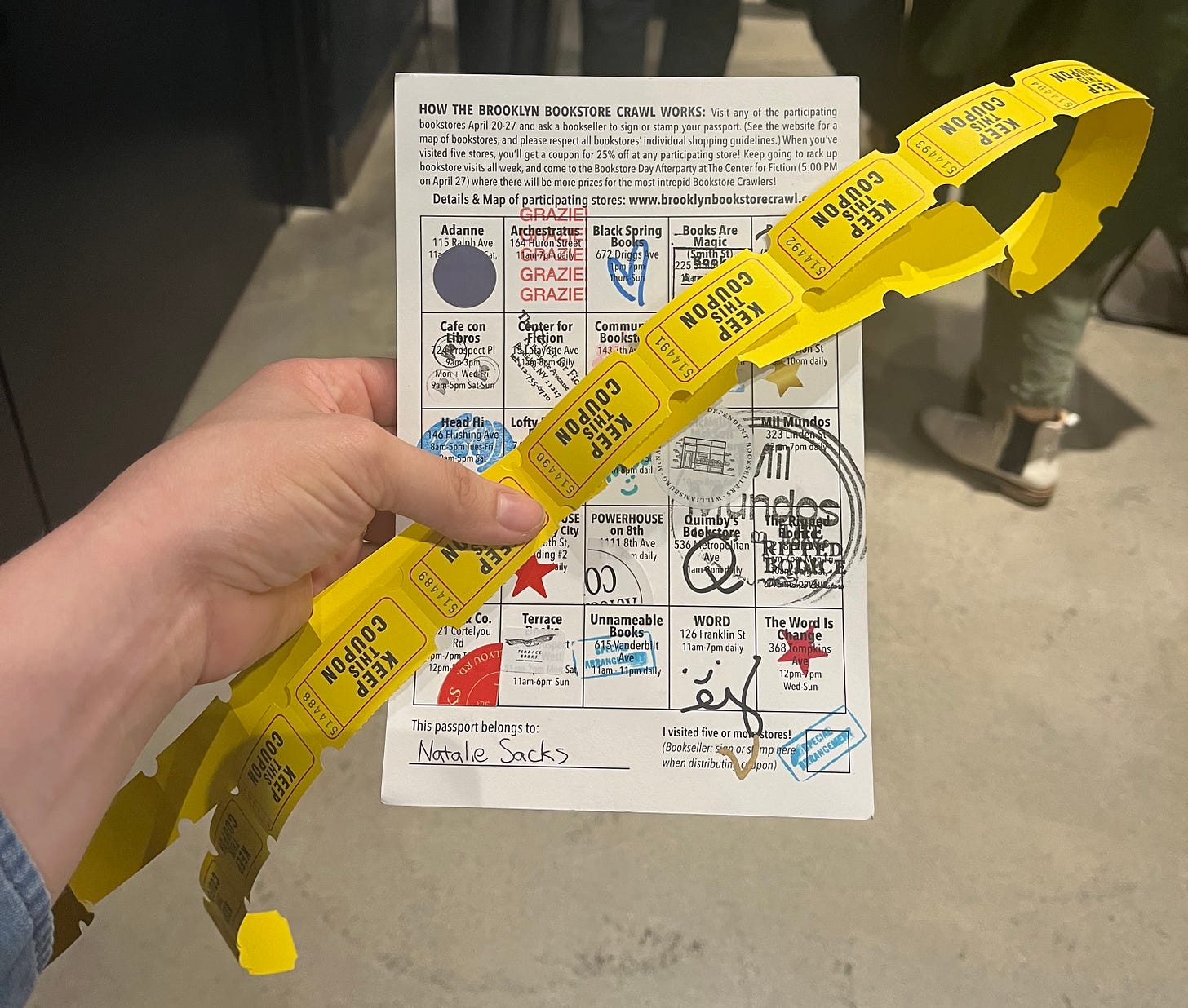 A card with 25 bookstores listed on it in squares. Each square is stamped or stickered. A hand holds a long line of raffle tickets in front of the card. A card with 25 bookstores listed on it in squares. Each square is stamped or stickered. A hand holds a long line of raffle tickets in front of the card.
