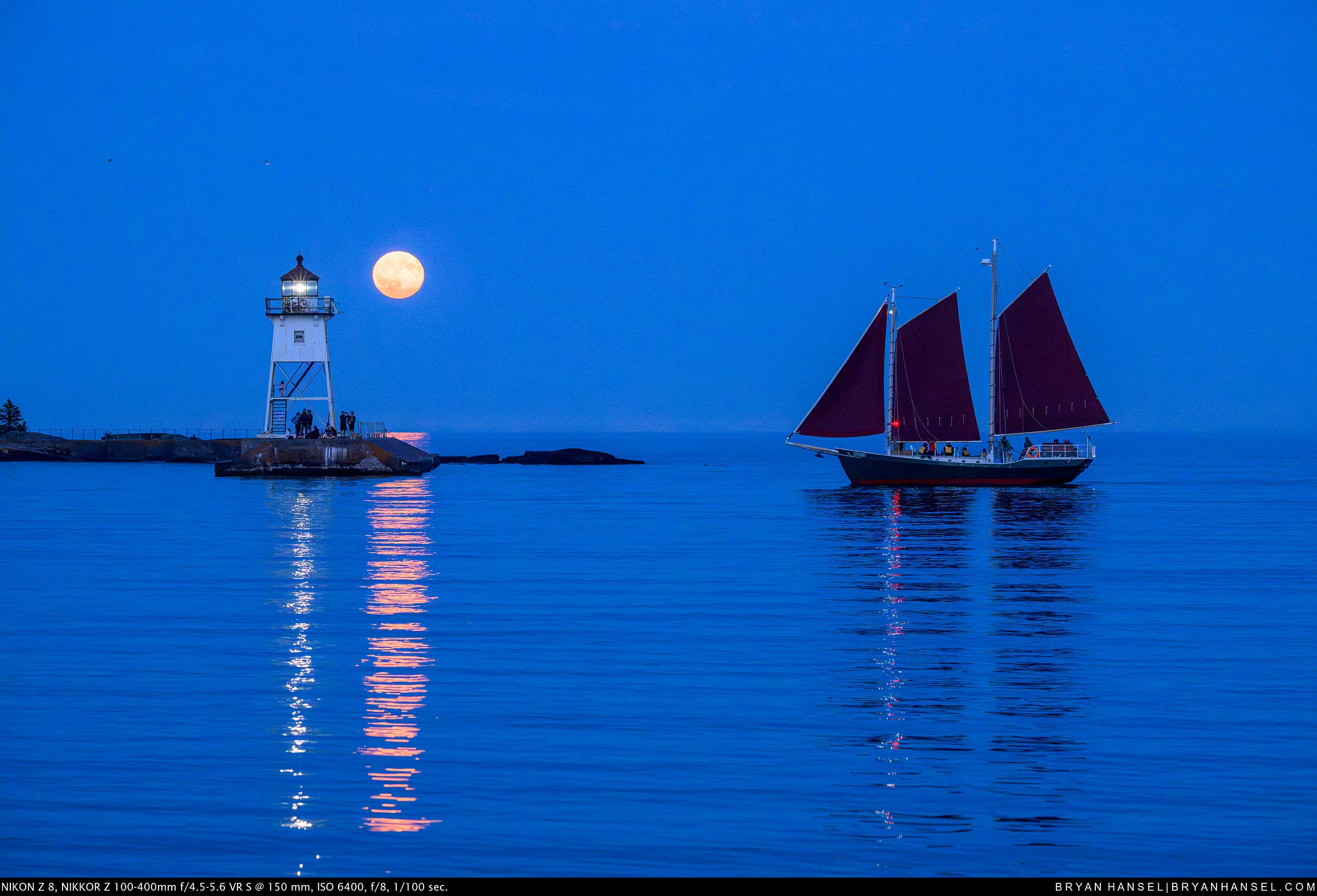 The nearly full moon rises beside the Grand Marais lighthouse as the schooner Hjørdis sails across the harbor beneath a deep blue evening sky.