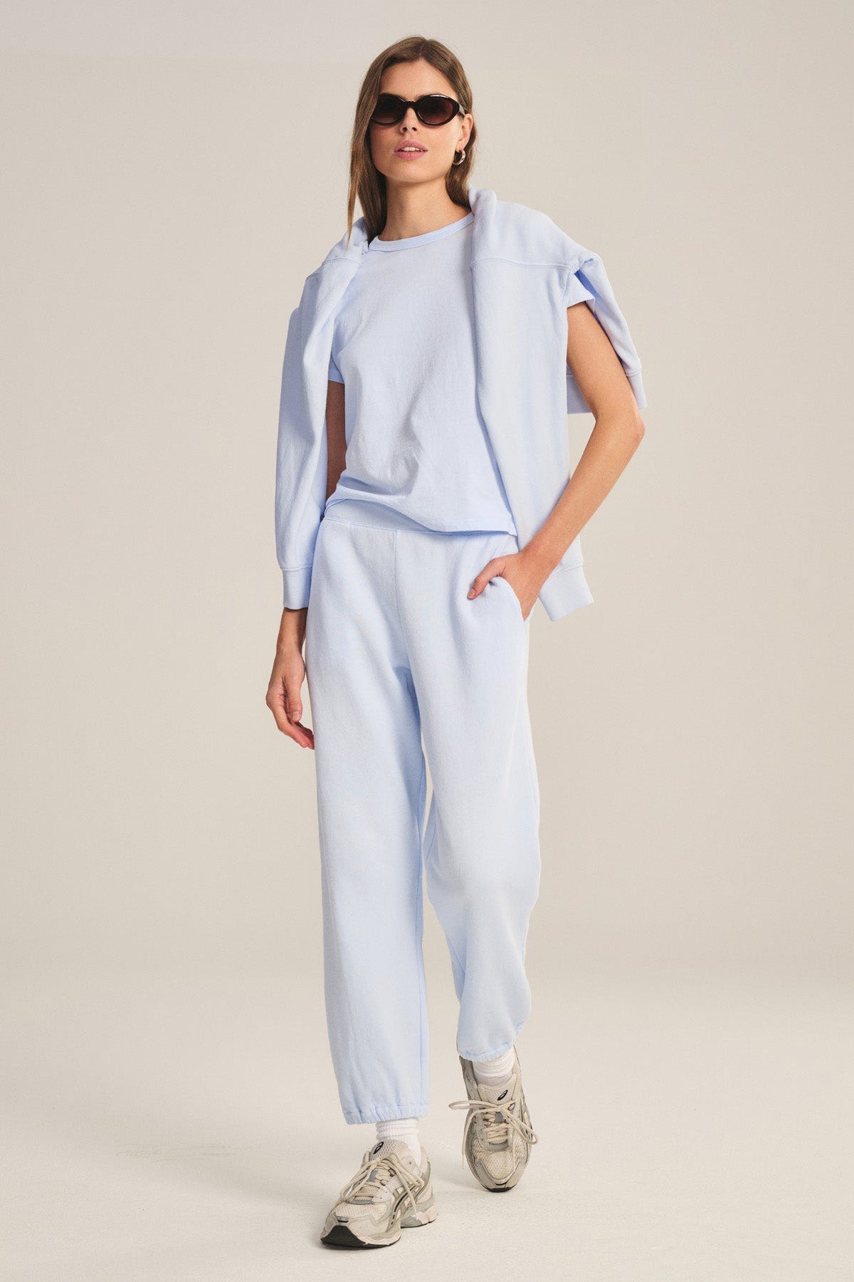 A person stands in a neutral studio wearing light blue ZARA SWEATPANT by Velvet by Graham & Spencer, matching joggers, sneakers, a relaxed fit sweatshirt draped over their shoulders, and sunglasses.